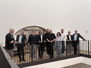 Gruppe von zehn Personen steht auf einem Balkon mit schwarzem Geländer in einem modernen Innenraum