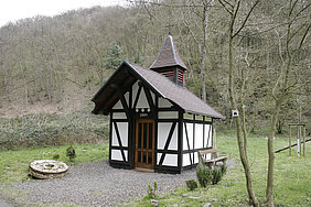 Kleine Fachwerkkapelle mit Holztür und spitzem Dach in einer bewaldeten Landschaft