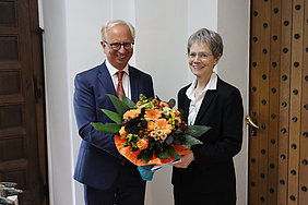 Zwei ältere Personen in formeller Kleidung, der Mann überreicht der Frau einen großen Blumenstrauß mit orangefarbenen und gelben Blüten