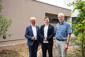 Drei Männer in formeller und legerer Kleidung stehen vor einem Gebäude bei einer Spendenübergabe