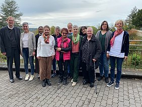 Gruppe von zwölf älteren und mittleren Erwachsenen, die draußen auf gepflastertem Weg stehen, im Hintergrund Bäume und ein Fluss, Anlass Jahrestag Senioren Seelsorge